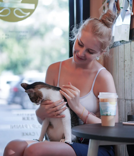 A woman in a white tank top smiles while gently petting a cat in her arms at a cozy café. A takeout coffee sits on the table nearby.